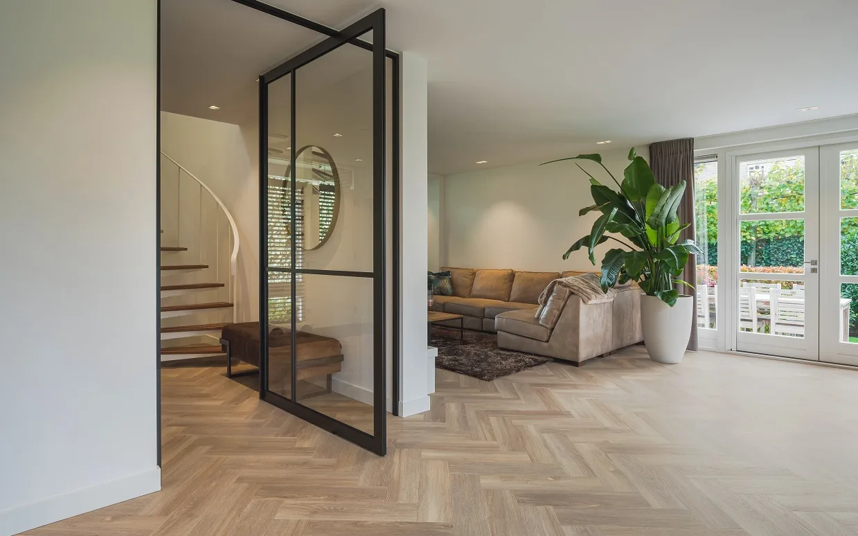 Moderne woonkamer met visgraat vloer, beige hoekbank en zwarte stalen deur.