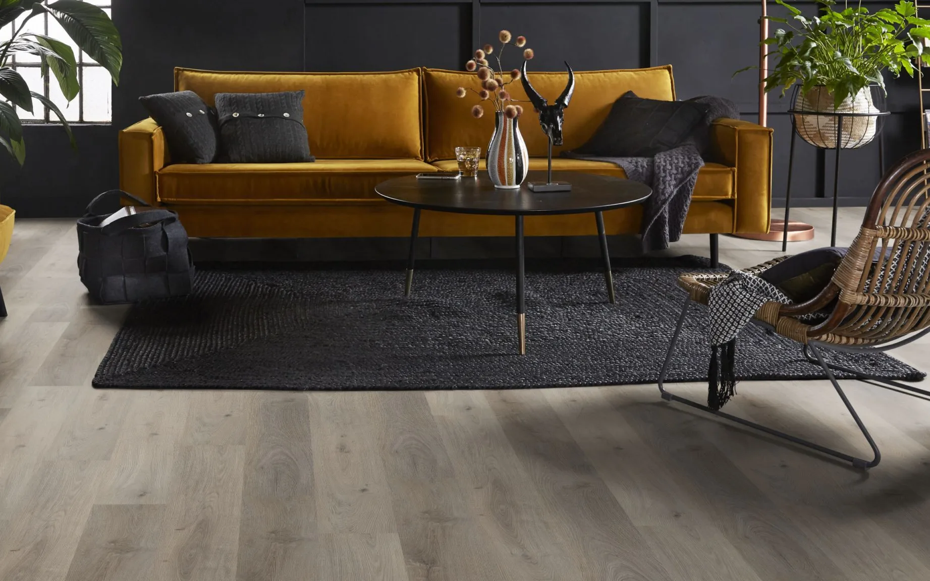 Woonkamer met vinylvloer in vergrijsde bruintint, oker kleurige bank en zacht vloerkleed.