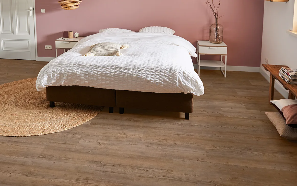 Slaapkamer met donkerbruine houtvloer, tweepersoonsbed en roze behangpapier.