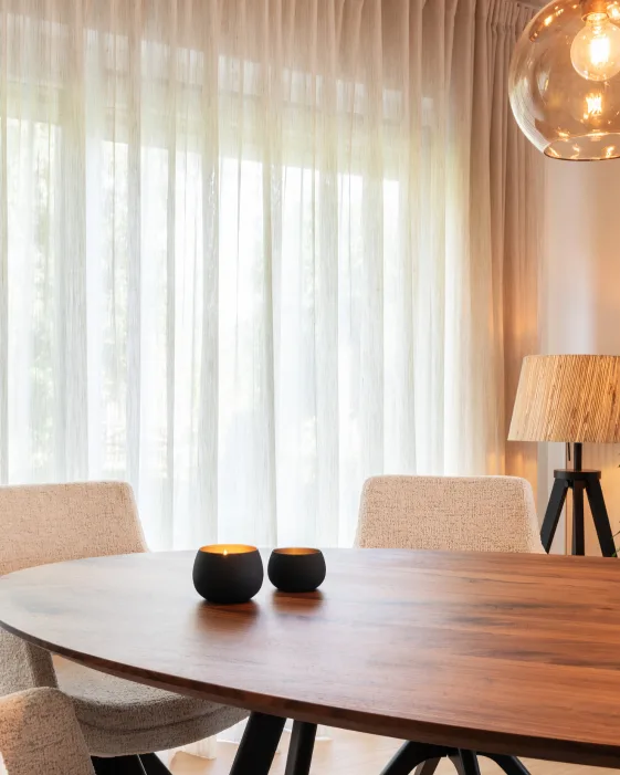 Eetkamer met licht transparante, kamerhoge gordijnen en een ovale houten eettafel met roomwitte stoelen