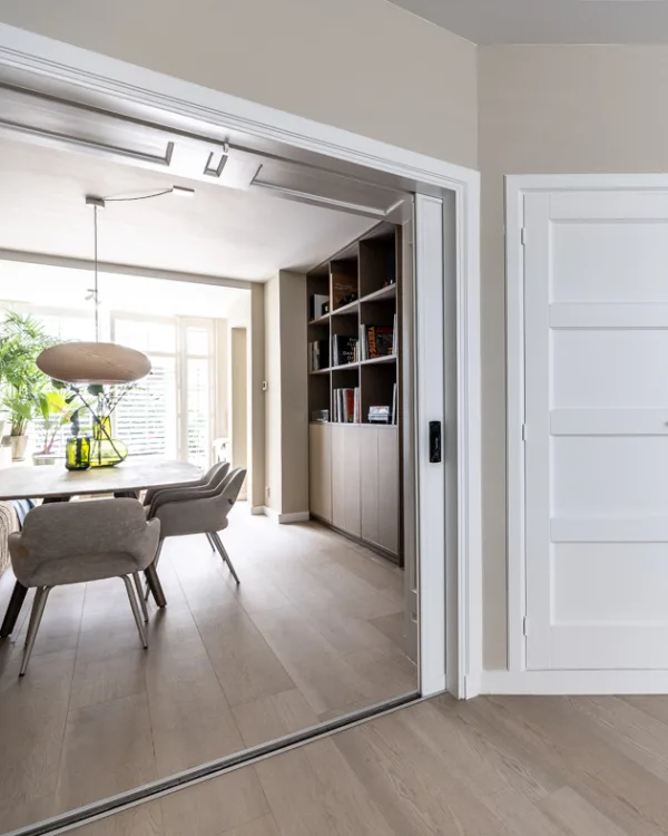 Woonkamer met grote tafel, boekenkast, ensuite deuren en vinylvloer in lichtbruin.