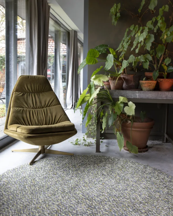Woonkamer met fluwelen relaxfauteuil, grijze gordijnen, planten en rond gemêleerd vloerkleed van wol.